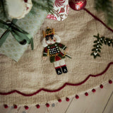 A festive Christmas tree skirt made of beige burlap fabric with a scalloped dark red border and pom-pom trim, decorated with a felt nutcracker and green pine branch accents. The skirt is styled under a Christmas tree with wrapped presents on a light wood floor.