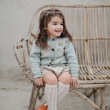 Little cute girl sitting in chair wearing hand knitted cardigan in duck blue which has beautifully hand embroidered floral design in the front part, it suits perfectly with her outfit
