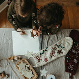 Two children in a festive setting with a few different decorations and products, including the cream white Gingerbread Stocking, showcasing their holiday charm and various designs.