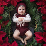 Baby in a crib wearing a dark grape romper and matching snowflake bonnet, surrounded by Christmas poinsettias in a festive ambient.