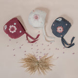 flat lay picture of 3 different coloured piece of hand knitted bonnet with string ties on it and with floral embroidery on both sides of the bonnet