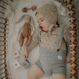 cute picture of seeping baby boy with his doll  wearing hand knitted sweater oats and bonnet in bubble style and hand knitted suspender shorts in duck blue