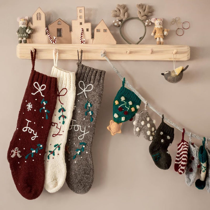 Cream white Gingerbread Stocking displayed on a wall-mounted hook with other stockings and festive decorations, highlighting its knitted texture and hand-embroidered JOY/snowflake pattern.