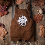 Caramel-colored hand-knitted snowflake romper displayed on a table, surrounded by dry leaves and a wooden mouse toy, emphasizing its large embroidered snowflake and soft merino wool.