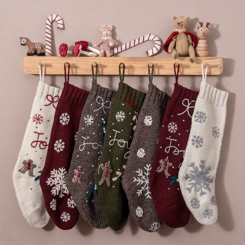 Various hand-knitted Christmas stockings in multiple colors, including Cream White, hanging in a festive holiday setting, showcasing their unique hand-embroidered patterns.