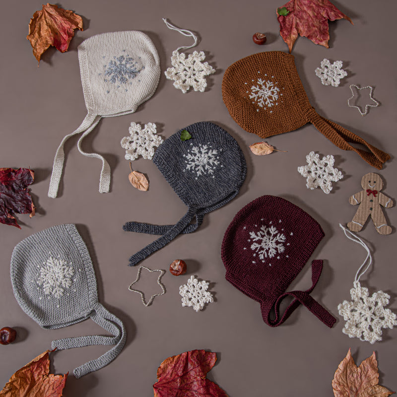 Collection of hand-knitted snowflake bonnets in various colors, including caramel and other shades, showcasing the embroidered snowflakes and tie bands.