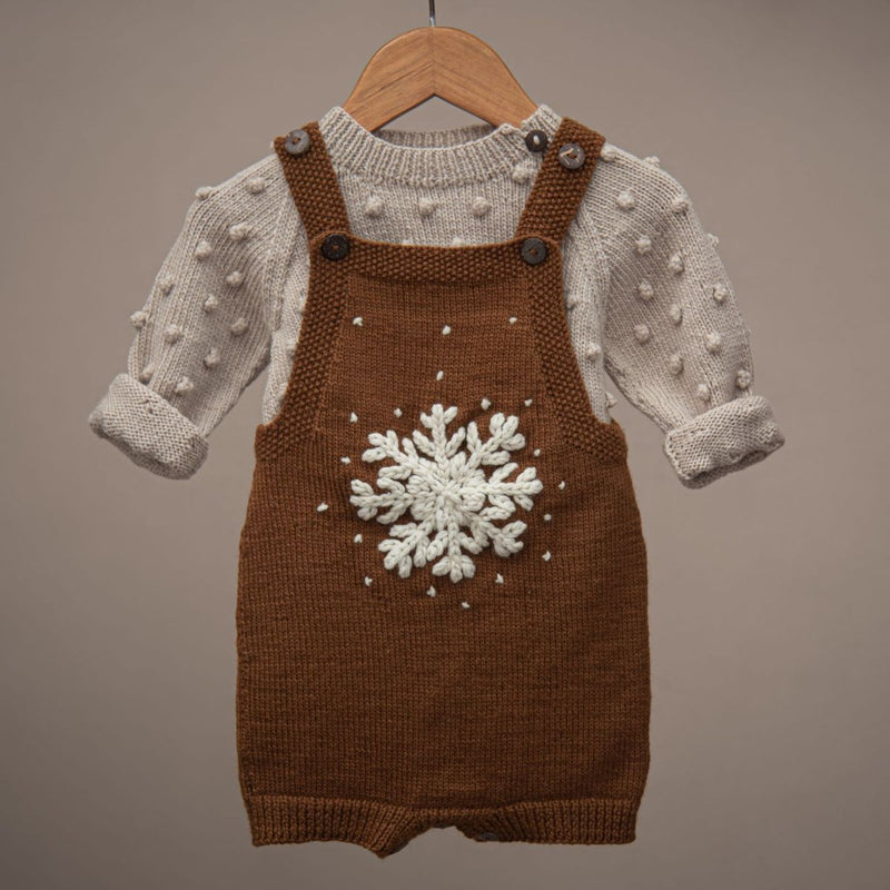 Caramel-colored hand-knitted snowflake romper draped over a sweater on a hanger, highlighting the large embroidered snowflake and soft merino wool.