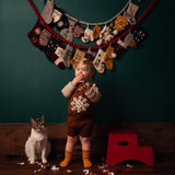 Child wearing a caramel-colored hand-knitted snowflake romper, with a cat and wooden red steps nearby. In the background, colorful stockings are hanging on the wall, adding a festive touch to the scene.
