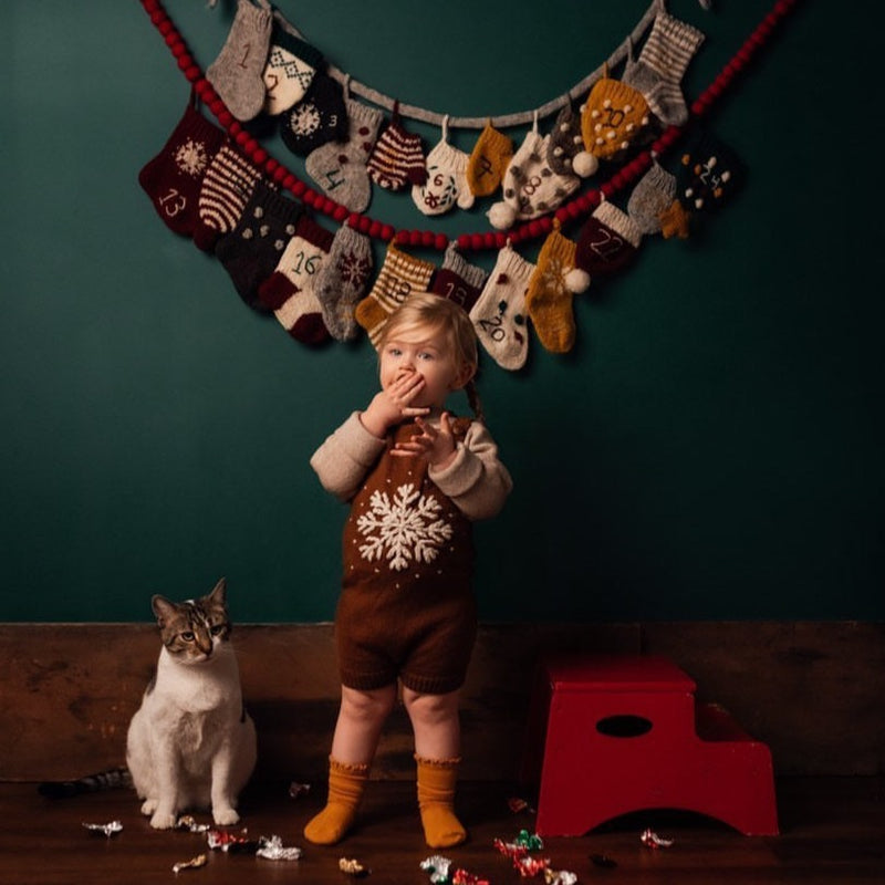 Child wearing a caramel-colored hand-knitted snowflake romper, with a cat and wooden red steps nearby. In the background, colorful stockings are hanging on the wall, adding a festive touch to the scene.