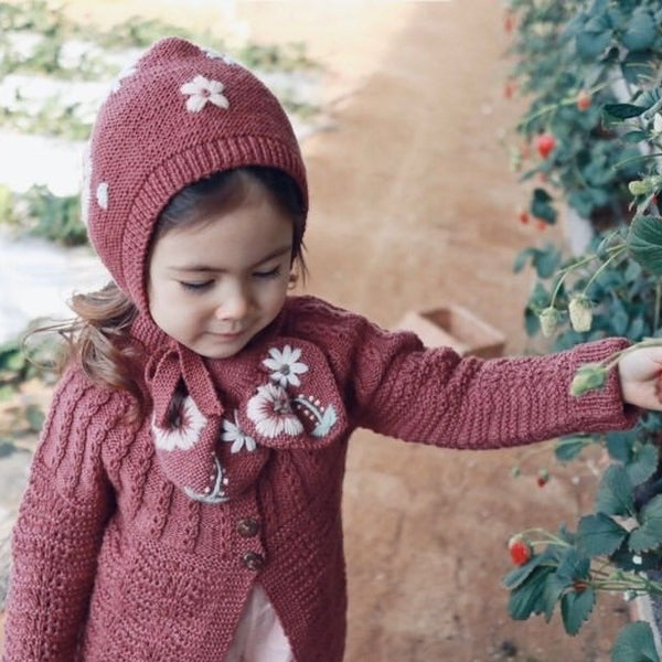 Cute little girl wearing hand knitted cardigan with hand knitted bonnet on deep berry which has floral embroidery on it