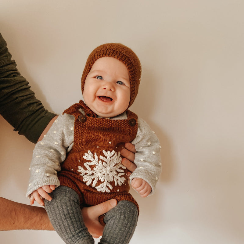 Baby wearing a caramel-colored hand-knitted snowflake romper, paired with rib tights, a knitted bonnet, and a sweater, showcasing the detailed embroidery and cozy winter outfit.