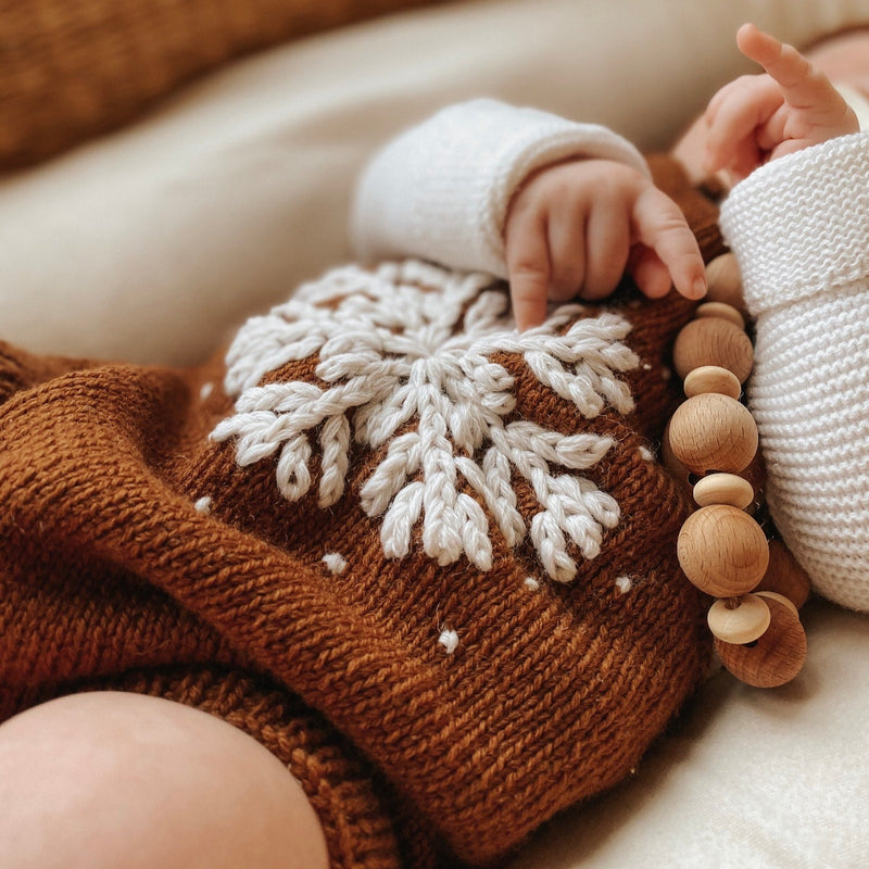 Close-up of a baby wearing a caramel-colored hand-knitted snowflake romper, featuring detailed embroidery of a large snowflake and scattered snow, showcasing the soft merino wool texture and scattered snow details.